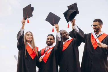 Bir grup farklı mezun, başarılarını havada şapkalarla kutluyorlar. Gülümseyen ve diploma taşıyan insanlar, neşe ve başarı gösterirler..