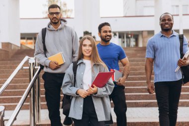 Sırt çantaları ve kitaplar taşıyan, arkadaşlıklarını ve akademik yaşamlarını sergileyen, gülümseyen ve kampüs merdivenlerinden inen dört üniversite öğrencisinden oluşan çeşitli bir grup..