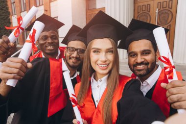 Başlık ve cüppeli çeşitli mezunlar başarılarını diplomalarla kutluyor, okul binasının önünde neşe ve başarılarını dile getiriyorlar..