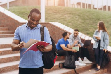 Afro-Amerikalı özgüvenli öğrenci kitap tutuyor ve arkadaşlarıyla kampüs merdivenlerinde gülümsüyor. Üniversitede arkadaşlığın ve akademik hayatın özünü yakalamak..