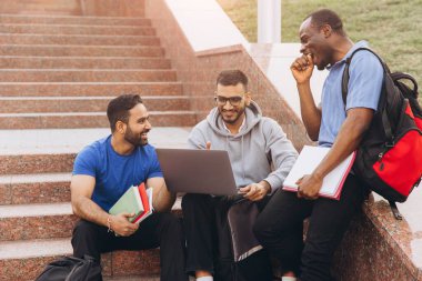 Üç farklı öğrenci kampüs basamaklarında oturmuş dışarıda ders çalışırken ve takım çalışması yaparken mutlu bir şekilde iş birliği yapıyorlar..