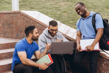 Bir grup kolej öğrencisi dışarıdaki merdivenlerde oturuyor, çalışmak için dizüstü bilgisayar kullanıyor ve gülüyorlar..