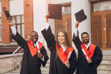 Üç farklı mezun dışarıda başarılarını kutluyor, ellerinde şapkalar ve diplomalar var. Onların ifadeleri mezuniyet gününde neşe ve başarıyı yansıtır..