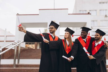 Akademik şapkalar ve elbiseler giyen, diplomalarını tutan, neşe ve başarılarını ifade eden çeşitli gülümseyen mezunlardan oluşan bir grup başarılarını dışarıda kutluyor..