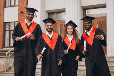 Dört mutlu mezundan oluşan çeşitli bir grup, bir üniversite binasının önünde diplomalarını tutarak kep ve cübbe giymedeki başarılarını kutluyor..