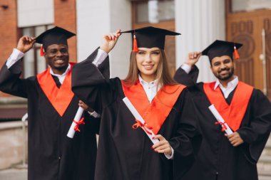 Çeşitli mezunlardan oluşan neşeli bir grup, başarılarını dışarıda kutluyor, ellerinde diplomalar ve kırmızı aksanlı geleneksel kepler ve elbiseler var..