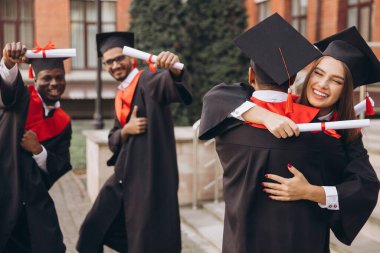 Çeşitli mezunlardan oluşan neşeli bir grup kep ve cübbe başarılarını kutluyor, diploma tutuyor ve birbirlerine sarılıyorlar. Bir üniversite binasının önünde yakalanmış..