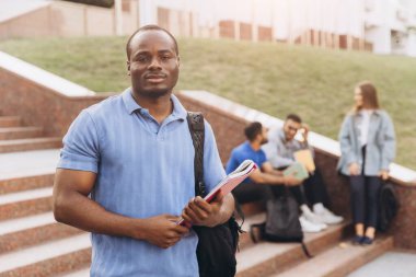 Afro-Amerikalı özgüvenli öğrenci kitap tutuyor ve arkadaşlarıyla kampüs merdivenlerinde gülümsüyor. Üniversitede arkadaşlığın ve akademik hayatın özünü yakalamak..