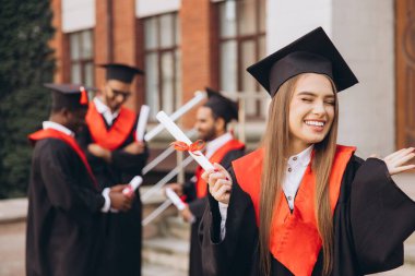 Mutlu genç bir kadın arkadaşlarıyla birlikte mezun oluyor, akademik elbiseler ve havan mermileriyle dışarıda kutlama yapıyor, özel bir günde neşe ve başarıyı ifade ediyor..