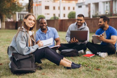 Mutlu ve çeşitli üniversite öğrencileri kampüste birlikte okuyorlar. Dışarıda dizüstü bilgisayarlar ve dizüstü bilgisayarlarla toplanıyorlar, rahat bir öğrenme ortamında takım çalışmalarını ve arkadaşlığı teşvik ediyorlar..