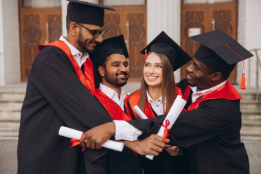 Bir grup farklı mezun, diplomalarını tutarken geleneksel kep ve cübbeler giyip, başarılarını dışarıda kutlamaktan mutluluk duyuyorlar..