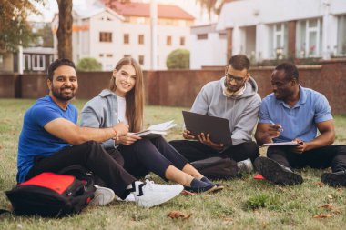 Genç üniversite öğrencileri kampüsün bahçesinde oturup bir çalışma projesinde işbirliği yapıyorlar. Bağlı ve odaklanmış halde fikirleri paylaşıyor ve doğal bir ortamda birlikte çalışıyorlar..