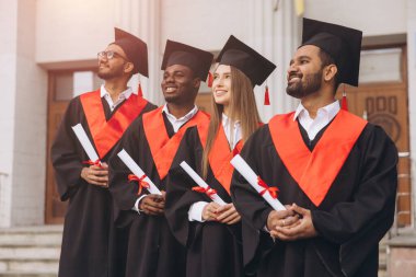 Başlık ve cübbe giyen çeşitli mezunlar, başarılarını dışarıda gülümseyerek ve neşeyle kutluyorlar..