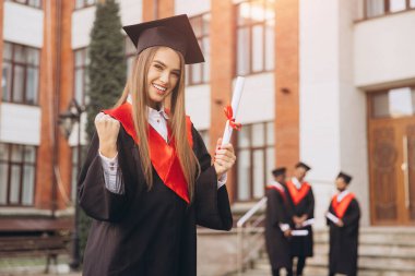 Mezuniyet şapkası ve cübbesiyle gülümseyen genç bir kadın, elinde diplomasıyla, başarıyı ve başarıyı sembolize ediyor, arka planda güneşli bir kampüs gününde sınıf arkadaşları var..