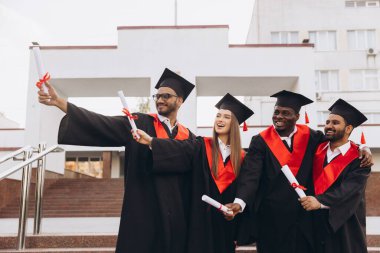 Akademik şapkalar ve elbiseler giyen, diplomalarını tutan, neşe ve başarılarını ifade eden çeşitli gülümseyen mezunlardan oluşan bir grup başarılarını dışarıda kutluyor..