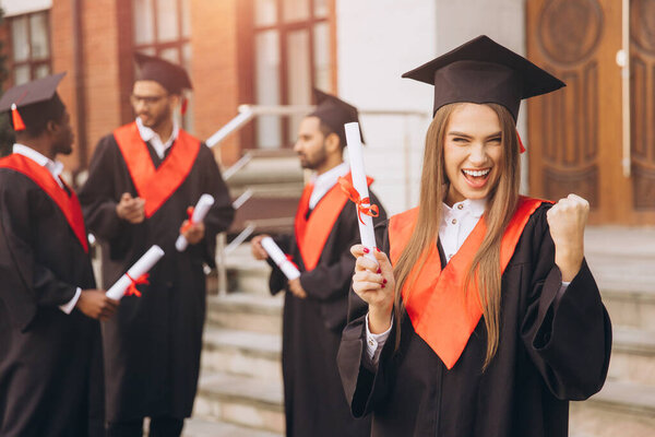 Счастливая молодая женщина выпускников с друзьями, празднуя на открытом воздухе в академических платьях и минометов, выражая радость и успех в особый день.