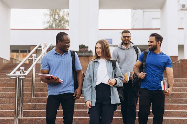Разнообразная группа студентов, идущих по ступеням кампуса, улыбается и разговаривает. Они носят с собой книги и рюкзаки, наслаждаясь приятным днем вместе.