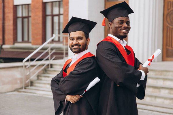 Two graduates in caps and gowns stand back-to-back, holding diplomas and smiling. A celebration of academic achievement at a university campus.