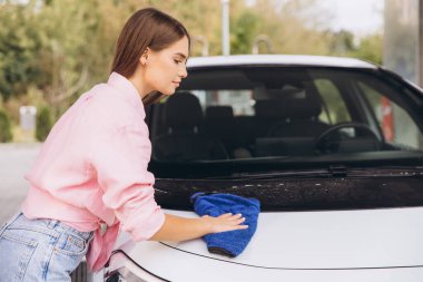 A young woman wipes the car hood with a blue cloth at an outdoor car wash. She wears a pink shirt and jeans, focusing on detail and cleanliness. clipart