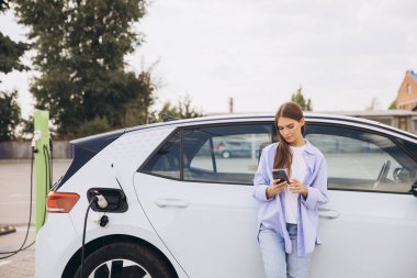 Genç bir kadın şarj istasyonunda elektrikli arabasına yaslanır, akıllı telefonuyla meşgul olur. Sahnede modern teknoloji, sürdürülebilirlik ve elektrikli araçların kolaylığı vurgulanıyor.