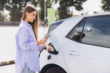 Genç Kadın, güneşli bir günde dışarıda akıllı telefon kullanırken elektrikli arabayı şarj ediyor, Yeşil Enerji ve Sürdürülebilir Yaşam tarzını teşvik ediyor