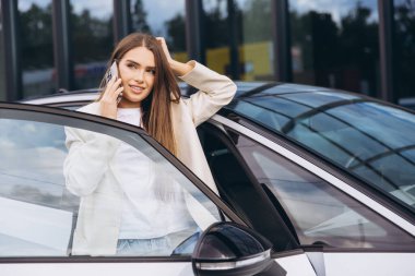 Kendine güvenen genç bir kadın arabanın kapısına dayanmış, akıllı telefonuyla konuşuyor. Beyaz ceketli sıradan bir kıyafet giyiyor. Profesyonel ama rahat bir tavır sergiliyor. Modern kentsel ortam.