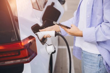 Bir kadın elektrikli arabasını akıllı telefon kullanarak şarj ediyor, modern çevre dostu teknolojiyi yansıtıyor ve kentsel bir ortamda sürdürülebilir yaşam sürüyor..
