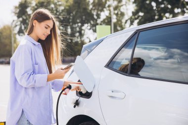 Genç Kadın, güneşli bir günde dışarıda akıllı telefon kullanırken elektrikli arabayı şarj ediyor, Yeşil Enerji ve Sürdürülebilir Yaşam tarzını teşvik ediyor