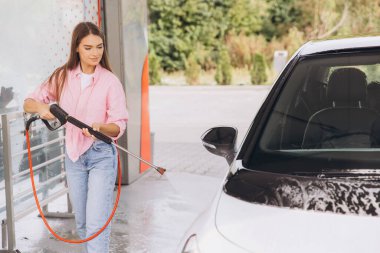 A young woman is cleaning her car with a pressure washer at a self-service car wash. She appears focused and content while effectively managing the washing process. clipart