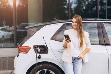 Genç kadın elektrikli arabanın yanında duruyor, şarj olurken akıllı telefonunu kontrol ediyor. Sıradan giyinmiş, modern elektrikli araç teknolojisinin keyfini çıkarıyor..