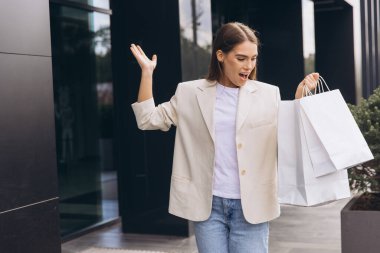 Neşeli bir kadın modern bir binanın önünde dururken birden fazla alışveriş çantası tutuyor. Yüzündeki ifade heyecan ve memnuniyeti yansıtıyor. Başarılı bir alışveriş gezisinin özünü yakalıyor..