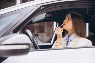 Bir kadın arabasında otururken ruj sürüyor. Açık renkli bir ceket giyiyor ve odaklanmışa benziyor..