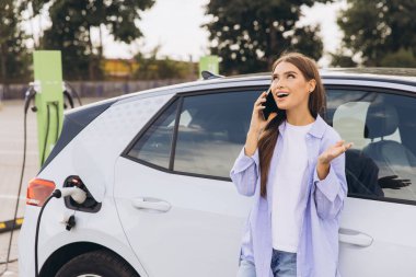 Genç kadın elektrikli arabanın yanında duruyor, arabası istasyonda şarj olurken cep telefonuyla konuşuyor, modern teknolojiyi ve çevre dostu ulaşımı simgeliyor..