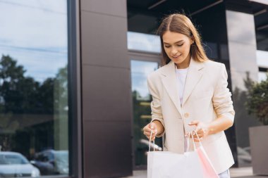 Alışveriş torbalarıyla genç bir kadın, modern bir şehir dükkanının önünde yürürken gülümsüyor. Şık bir ceket giymiş, neşe ve moda anlayışı yayıyor. Yaşam tarzı ve alışveriş temaları için ideal görüntü.