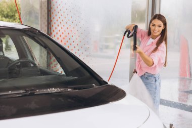 Gülümseyen genç bir kadın otomobil yıkamada yüksek basınçlı hortumla arabasını temizliyor. Neşeli ve odaklanmış görünüyor, modern araba yıkama hizmetlerinin kolaylığını gösteriyor.