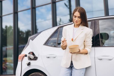 Genç bir kadın şarj istasyonunda elektrikli bir arabaya yaslanırken sağlıklı bir salatanın tadını çıkarıyor. Sahne sürdürülebilirlik, modern yaşam tarzı ve kolaylık sunar..