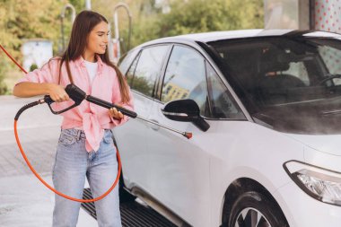 Genç bir kadın açık hava oto yıkama makinesinde arabasını yıkamaktan hoşlanıyor. Gülümsüyor ve günlük giysiler giyiyor, temizlik sırasında neşeli bir atmosfer yaratıyor.