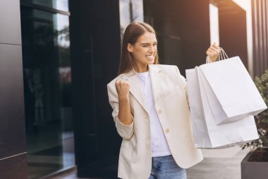 Neşeli bir kadın elinde alışveriş torbalarıyla dışarıda duruyor ve başarılı alışveriş deneyimini kutluyor. Heyecan ve memnuniyeti ifade ediyor, olumlu bir perakende terapi anını gösteriyor..