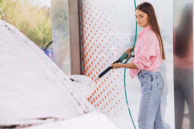 A young woman cleans her car at a self-service car wash using a hose. She is focused on washing the vehicle, surrounded by soap suds and sunlight. clipart