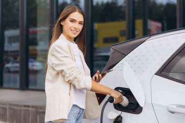 Genç bir kadın modern bir şarj istasyonunda elektrikli arabasını şarj ediyor. Telefonunu tutarken güvenle gülümsüyor. Çevre dostu ve sürdürülebilir enerji seçeneklerini vurguluyor..