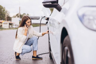 Genç bir kadın elektrikli arabasının yanında çömelmiş şarj olurken ve telefonuyla konuşurken. Kentsel arka plan modern şehir yaşam tarzını ve çevre dostu ulaşım seçeneğini gösteriyor. Arabayla ilgili sorun.