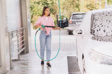 Pembe gömlekli genç bir kadın self-servis istasyonunda arabasını yıkıyor. Köpükle çevrili beyaz aracı temizlemek için yüksek basınçlı hortum kullanıyor..