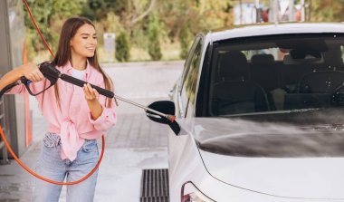 Genç bir kadın açık hava oto yıkama makinesinde arabasını yıkamaktan hoşlanıyor. Gülümsüyor ve günlük giysiler giyiyor, temizlik sırasında neşeli bir atmosfer yaratıyor.