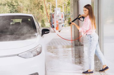 Genç bir kadın yüksek basınçlı hortumla arabasını oto yıkamada zevkle yıkıyor. Aydınlatma güneşli bir günde açık havada, temizliği ve özeni vurguluyor..