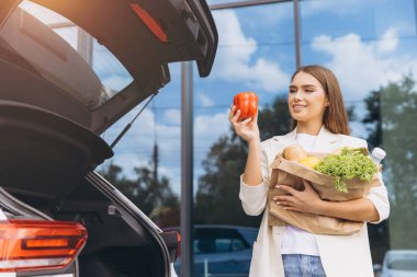 Bir kadın bagajından alışveriş yaparken elinde kırmızı bir dolma biber tutarken gülümsüyor. Taze ürünlerle dolu bir kese kağıdı taşıyor. Sahne alışverişin ve sağlıklı yaşamın neşesini yakalıyor..