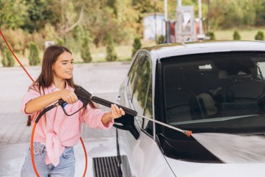 Genç bir kadın açık hava oto yıkama makinesinde arabasını yıkamaktan hoşlanıyor. Gülümsüyor ve günlük giysiler giyiyor, temizlik sırasında neşeli bir atmosfer yaratıyor.