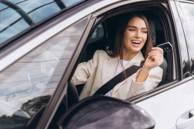 Neşeli bir kadın arabasında oturuyor, elinde bir anahtar var ve gülümsüyor, yeni araba alımını kutluyor. Görüntü mutluluk, özgürlük ve başarıyı iletir..