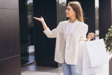 Bej ceketli genç bir kadın alışveriş torbaları tutuyor ve modern bir binanın önünde el kol hareketi yapıyor. Neşeli görünüyor, başarılı bir perakende terapi ve şık moda gününü kucaklıyor..