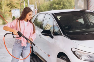 Genç bir kadın açık hava oto yıkama makinesinde arabasını yıkamaktan hoşlanıyor. Gülümsüyor ve günlük giysiler giyiyor, temizlik sırasında neşeli bir atmosfer yaratıyor.