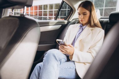 Bej ceketli ve kot pantolonlu neşeli bir kadın arabada otururken akıllı telefonuyla video görüşmesi yapıyor. Keyfi yerinde ve sohbete dalmış görünüyor..